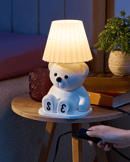 White bear-shaped lamp with pleated shade on a wooden table, plugged into a power source.