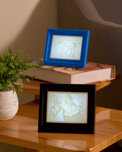 Two digital photo frames, one blue and one black, displaying images on a wooden surface with a plant and book in the background.